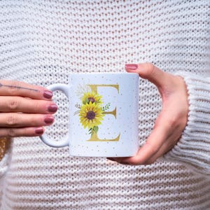 Pode incluir: Caneca de cerâmica branca com a letra dourada "E" e um desenho de girassol. A caneca tem uma alça branca e é decorada com pequenos pontos dourados. A caneca é segurada por uma pessoa que usa um suéter branco.