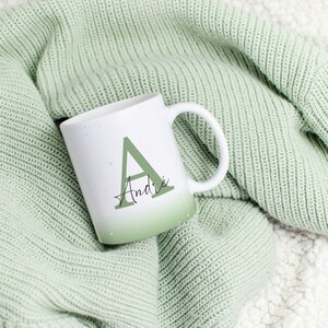 May include: A white ceramic mug with a green ombre design, displaying the letter "A" and the name "Andre" in a decorative font. The mug is placed on a soft, textured green blanket, creating a warm and inviting atmosphere.