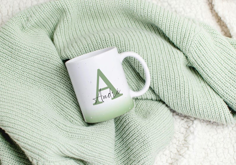 May include: A white ceramic mug with a green ombre design, displaying the letter "A" and the name "Andre" in a decorative font. The mug is placed on a soft, textured green blanket, creating a warm and inviting atmosphere.
