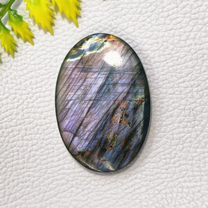 May include: Oval-shaped labradorite gemstone with a polished surface. The stone displays a mix of purple, blue, and gray hues with iridescent flashes. The gemstone has a smooth, glossy finish and is suitable for jewelry making.