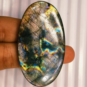 Peut inclure: Une pierre précieuse de labradorite de forme ovale présentant une surface polie et lisse avec des fractures internes visibles et une labradorescence vibrante affichant des éclats irisés de bleu, de jaune et d'or sur un fond gris foncé fumé.