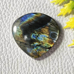 May include: A teardrop-shaped labradorite gemstone with iridescent blue, green, and gold flashes. The polished stone has a dark base color with streaks of color. The gemstone is set against a white textured background.