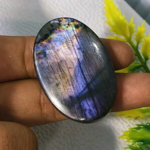 May include: An oval-shaped labradorite gemstone with a polished surface. The stone displays a mix of colors, including blues, purples, and hints of gold, with visible internal patterns. The gemstone is held in a hand.