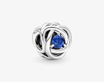 May include: A silver charm with a woven design, featuring a vibrant blue gemstone at its center. The charm is round and has a polished finish, creating a striking contrast between the metal and the gemstone. This jewelry piece is suitable for bracelets and necklaces.