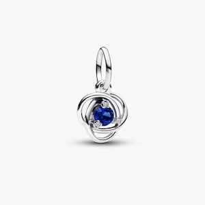 Könnte beinhalten: Ein silberfarbener Charm-Anhänger mit einem blauen Edelstein in der Mitte und zwei kleineren, klaren Steinen. Der Anhänger hat ein geschlungenes Design um den zentralen Stein und eine kleine Schlaufe oben zur Befestigung. Schmuck für ein Armband oder eine Halskette.