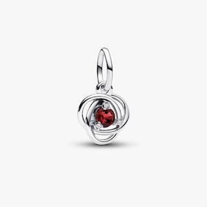 Puede incluir: Un charm plateado con una gema central de color rojo oscuro y forma ovalada. El charm presenta un diseño de nudo con tres pequeñas gemas transparentes que rodean la piedra central. Tiene un lazo en la parte superior para sujetarlo.