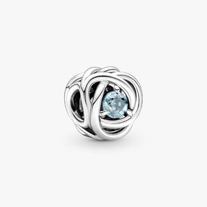 May include: A silver charm with a woven design, featuring a light blue gemstone at its center. The charm has a spherical shape and is set against a plain white background. The intricate metalwork surrounds the gemstone, creating a delicate and elegant appearance.