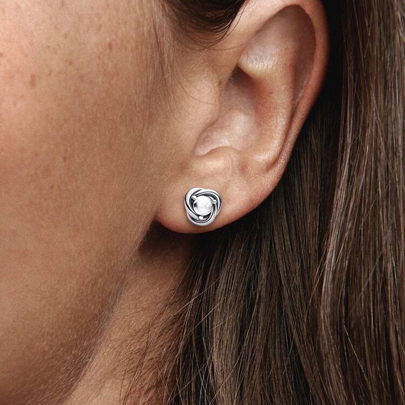 Puede incluir: Un pendiente de bot&oacute;n plateado con un dise&ntilde;o de nudo y una perla blanca central. El pendiente se lleva en una oreja, mostrando su dise&ntilde;o detallado y su apariencia elegante. Una joya cl&aacute;sica.