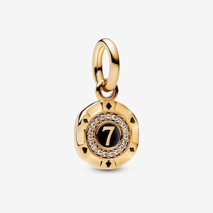 May include: A gold-colored charm with a round pendant featuring the number 7 in black, surrounded by clear crystals. The charm has a loop for attaching to a bracelet or necklace. The pendant is accented with small black shapes.