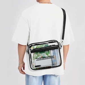 Peut inclure: Un sac bandoulière rectangulaire transparent avec des bordures noires et une sangle réglable noire. Le sac comporte une poche avant zippée et un compartiment principal contenant un insert rectangulaire blanc avec le texte YOUR DESIGN HERE.