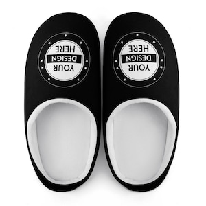 Pode incluir: Um par de chinelos pretos com interior branco. Cada chinelo apresenta um design circular com o texto "YOUR DESIGN HERE" em branco. Estes chinelos de interior est&atilde;o sobre um fundo branco.