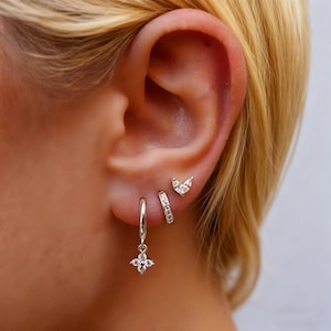 Puede incluir: Primer plano de una oreja adornada con tres pendientes diferentes. El primero es un aro plateado con una flor colgante de cuatro pétalos. El segundo es un pequeño aro plateado con cristales incrustados. El tercero es un pequeño pendiente plateado con forma de hoja.