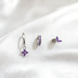 May include: A set of three silver earrings featuring purple gemstones. The set includes a hoop earring with purple stones, a hoop with a dangling flower, and a stud with a butterfly design.
