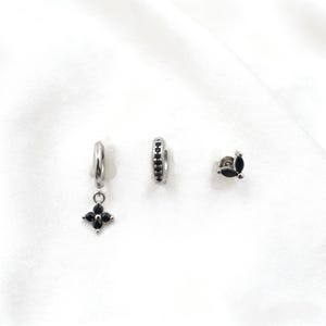 May include: A set of three silver-toned earrings. One earring has a black flower pendant, another is a small hoop with black stones, and the third is a stud with a cluster of black stones. The earrings are displayed on a white background.
