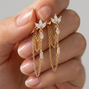 May include: A pair of gold dangle earrings featuring a cluster of three marquise-cut clear stones at the top, with delicate gold chains hanging down. Each chain ends in a small clear stone, with one teardrop-shaped and one rectangular-cut stone.