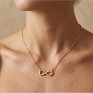May include: A gold-colored necklace featuring an infinity symbol pendant. The pendant is a continuous loop, suspended from a delicate chain. The necklace is worn around the neck, with the chain resting on the skin.