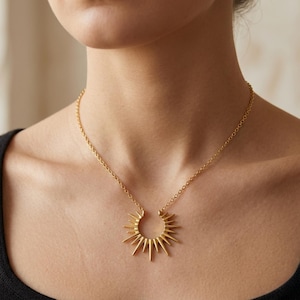 May include: A gold-colored necklace featuring a sunburst pendant. The pendant has a half-circle design with pointed rays extending outwards. The necklace is worn around the neck and hangs on a delicate chain.