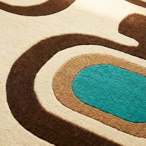 Puede incluir: Primer plano de una alfombra con dise&ntilde;o retro. La alfombra presenta un patr&oacute;n de l&iacute;neas curvas en tonos marr&oacute;n, beige, tostado y turquesa. La textura parece suave y afelpada, lo que sugiere un revestimiento de suelo c&oacute;modo.