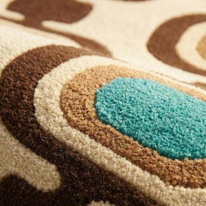 Puede incluir: Primer plano de una alfombra con dise&ntilde;o retro. La alfombra presenta formas curvas conc&eacute;ntricas en tonos marr&oacute;n, crema y turquesa. La textura parece suave y afelpada, con una pila alta. La est&eacute;tica general recuerda a la d&eacute;cada de 1970.