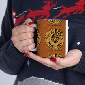 Puede incluir: Una taza de cerámica blanca con el texto "FOREVER DIVINE COFFEE" en un diseño circular con un gráfico de ángel. La taza tiene un fondo marrón con el texto "Mark Anthony GO Collection".