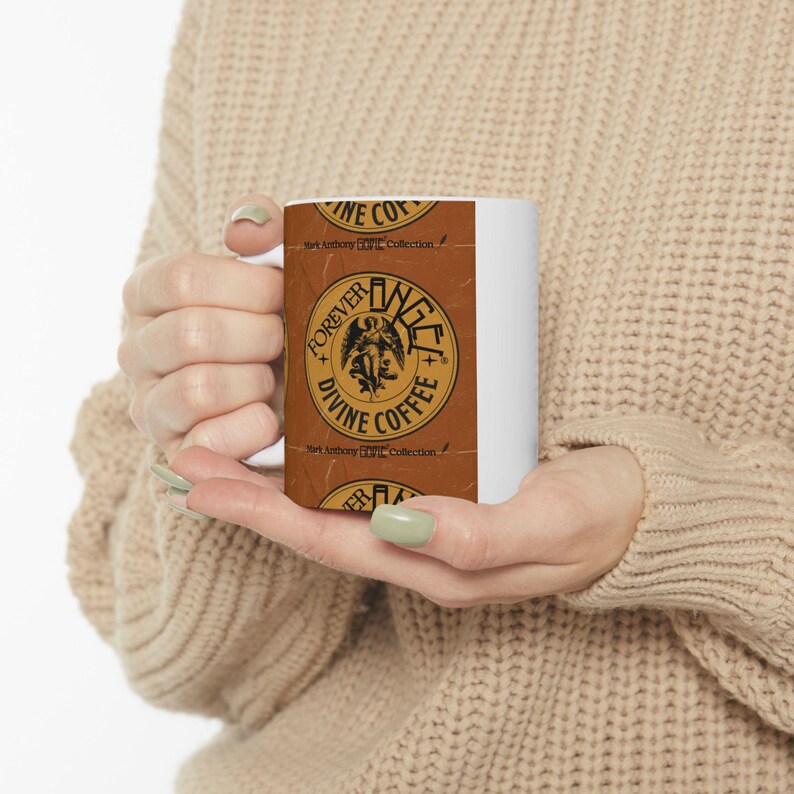 May include: A white ceramic coffee mug featuring a brown label with the words "FOREVER DIVINE COFFEE" in a circular design. The label also includes an angel illustration and the text "Mark Anthony Collection."