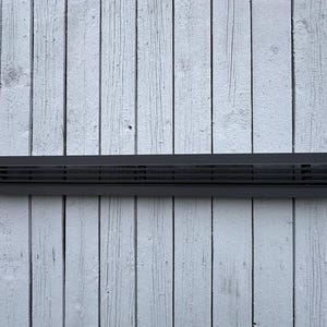 Może przedstawiać: Długa, prostokątna czarna osłona wentylacyjna z poziomymi listwami. Osłona wentylacyjna jest wykonana z metalu i ma matowe wykończenie. Jest zamontowana na zwietrzałej, białej drewnianej ścianie.