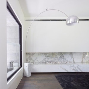 May include: A contemporary interior with a silver arc floor lamp and a spherical shade. The room features a marble accent wall, a dark rug, and hardwood flooring. A large window is visible on the left.