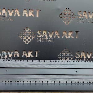 Może przedstawiać: Czarny metalowy szyld z wyciętym tekstem "SAVA ART METAL" w kilku miejscach. Poniżej prostokątny element metalowy o teksturowanej powierzchni i małych otworach na krawędziach. Szyld zamontowany na czarnej półce na tle ceglanej ściany.