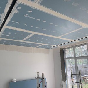 Puede incluir: Vista interior de una habitaci&oacute;n en construcci&oacute;n, con un techo parcialmente terminado con paneles de yeso azules y masilla blanca. Se instalan luminarias lineales negras. Se ve una ventana grande y una escalera.