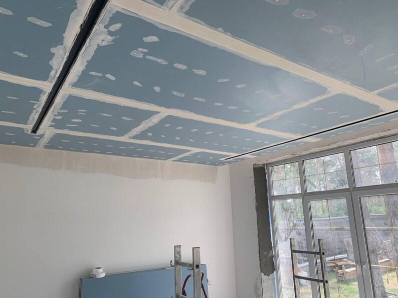 Puede incluir: Vista interior de una habitaci&oacute;n en construcci&oacute;n, con un techo parcialmente terminado con paneles de yeso azules y masilla blanca. Se instalan luminarias lineales negras. Se ve una ventana grande y una escalera.