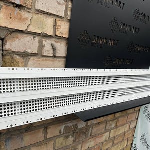 Może przedstawiać: Biały metalowy pasek wentylacyjny z wieloma rzędami okrągłych perforacji. Pasek jest umieszczony na tle ceglanej ściany i czarnego metalowego panelu z napisem "SAVA ART METAL".