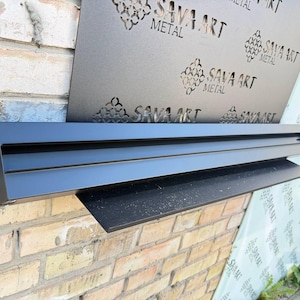 May include: A black metal shelf with a rectangular design is mounted on a brick wall. A decorative metal panel with the text "SAVA ART METAL" is visible behind the shelf. The shelf has a modern, minimalist aesthetic.