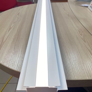 Puede incluir: Una luminaria lineal LED blanca, que emite luz brillante. La luminaria tiene forma rectangular con una tira de luz central y está colocada sobre un fondo de escritorio de madera. La luz está diseñada para uso en interiores.