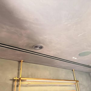 May include: A light grey ceiling featuring two black linear vents and recessed lighting. A gold-coloured metal rack is suspended below. The ceiling has a textured appearance, creating a modern and minimalist aesthetic.