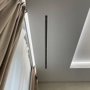 May include: A modern ceiling design with a long, rectangular air vent featuring a black mesh pattern. Beige curtains are on the left, and the ceiling is light grey. Recessed lighting is also visible in the design.