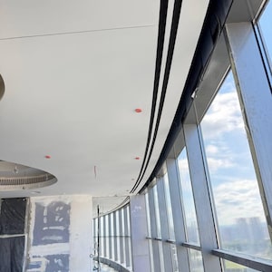 May include: Interior view of a modern building under construction. The image shows a curved wall of large windows, a white ceiling with black trim, and exposed construction materials. The sky is visible through the windows.