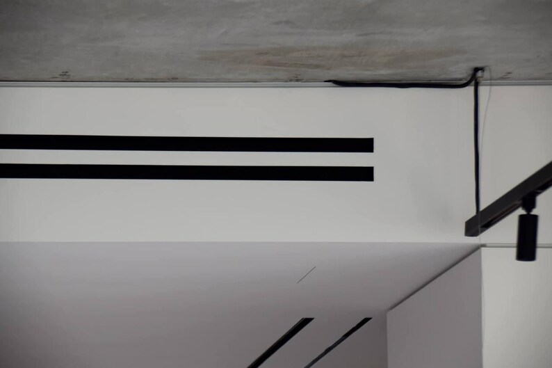 Puede incluir: Toma interior que muestra una pared blanca con dos franjas negras horizontales. Un cable negro recorre el techo y una l&aacute;mpara negra cuelga del techo. El techo es de hormig&oacute;n.