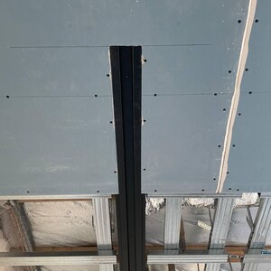 Puede incluir: Una escena de construcci&oacute;n que muestra una secci&oacute;n de techo con paneles de yeso y estructura met&aacute;lica. Una viga vertical de metal negro recorre el centro, con tornillos y costuras visibles. El techo es de color gris claro.