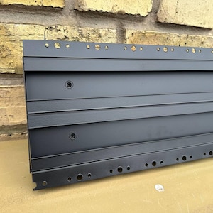 May include: A black metal panel featuring horizontal ridges and a series of holes along the top and bottom edges. The panel has circular indentations and is set against a brick wall background.