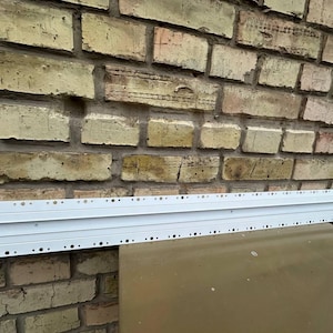 May include: A long, white metal strip with evenly spaced holes, possibly for mounting. It's set against a weathered brick wall. The strip is likely used in construction or a similar application. The length is approximately 100 cm.