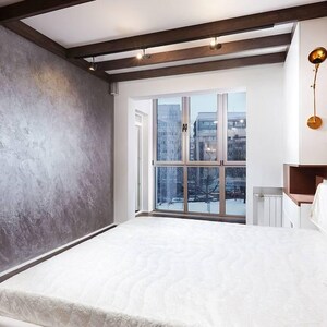 Puede incluir: Un dormitorio moderno con una gran ventana con vistas a un paisaje urbano. La habitaci&oacute;n cuenta con una pared gris texturizada, vigas de madera en el techo y una cama con un edred&oacute;n blanco. Un panel decorativo de madera est&aacute; detr&aacute;s de la cama.