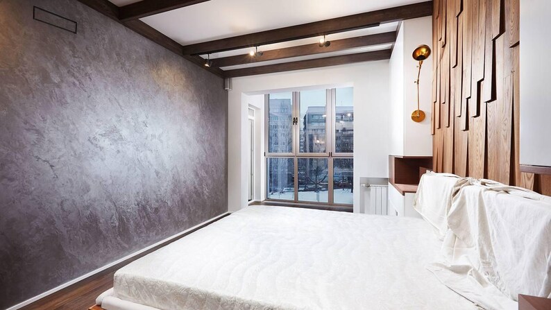 Puede incluir: Un dormitorio moderno con una gran ventana con vistas a un paisaje urbano. La habitaci&oacute;n cuenta con una pared gris texturizada, vigas de madera en el techo y una cama con un edred&oacute;n blanco. Un panel decorativo de madera est&aacute; detr&aacute;s de la cama.