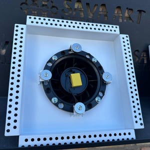 Peut inclure: Un cadre métallique carré blanc avec un ventilateur noir et un composant jaune au centre. Le cadre a une bordure perforée. Le fond présente le texte "SAVA ART" en couleur sombre.