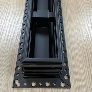 May include: A black metal vent cover with a rectangular frame and a series of small holes along the edges. The vent cover has a central recessed area and horizontal slats. The product is designed for air ventilation.