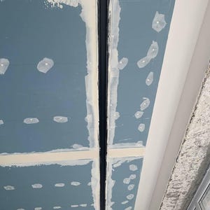 Puede incluir: Vista de cerca de un techo en construcci&oacute;n. El techo est&aacute; compuesto por paneles de yeso azules con masilla blanca y un canal empotrado negro. Se ven puntos blancos de masilla. Una pared blanca y un marco de ventana est&aacute;n en el fondo.