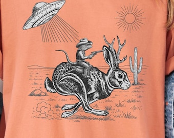 May include: Terracotta-colored t-shirt featuring a black and white graphic of a frog riding a jackalope, with a UFO and sun in the background. The shirt has the word "Terracotta" printed at the bottom.