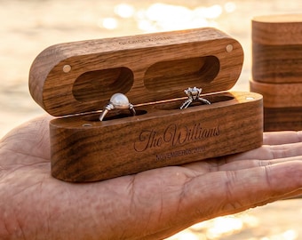 Könnte beinhalten: Eine rechteckige Ringbox aus Holz mit Klappdeckel, die zwei runde Innenfächer mit einem Perlenring und einem Diamantring enthält. Die Außenseite ist mit dem Text The Williams und NOVEMBER 2018 graviert, daneben stehen zwei weitere Boxen.