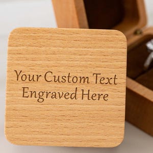 May include: A light-coloured wooden ring box, square in shape, with a hinged lid. The box is open, displaying a ring. The lid of the box has the text "Your Custom Text Engraved Here". The box is made of light-coloured wood.