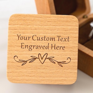 Op de afbeelding: Een vierkante houten ringdoos met een scharnierend deksel voorzien van de lasergegraveerde tekst "Your Custom Text Engraved Here" boven een hart- en bladerontwerp. De open doos toont een donkerbruine fluwelen binnenkant met een zilverkleurige ring.