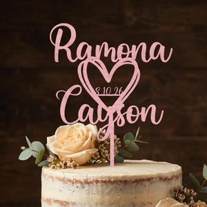 Op de afbeelding: Een lichtroze taarttopper met de namen Ramona en Cayson in een cursief lettertype, gescheiden door een hartvormige omtrek met de datum 8.10.26. De topper is geplaatst in een tweelaagse taart versierd met perzikkleurige rozen en eucalyptus.